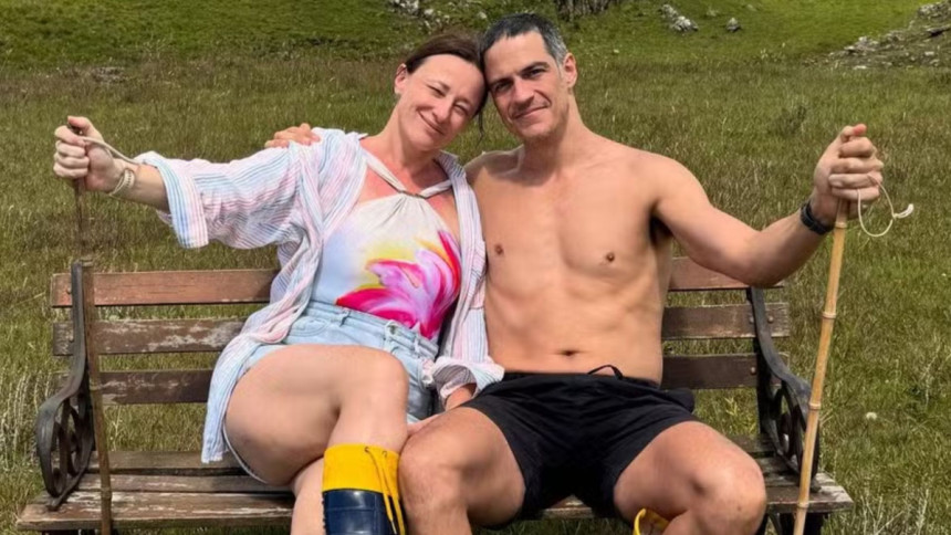 Mateus Solano sem camisa e Paula Braun com shorts jeans sorriem para a câmera, abraçados em um banco de madeira em meio à natureza.