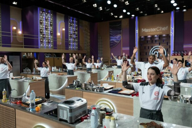 Os participantes do MasterChef Confeitaria em pé nas bancadas do estúdio, com as mãos levantadas para cima, celebrando a estreia do programa.