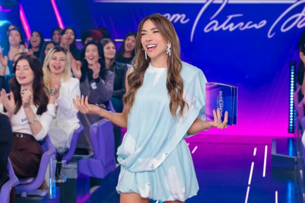 Apresentadora Patricia Abravanel, vestindo um vestido azul claro, sorri e gesticula no palco do Programa Silvio Santos, com a plateia ao fundo.