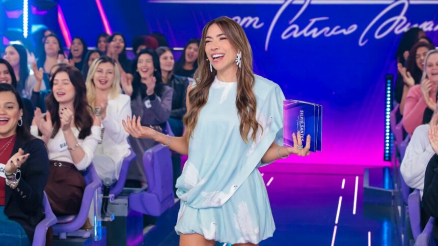 Apresentadora Patricia Abravanel, vestindo um vestido azul claro, sorri e gesticula no palco do Programa Silvio Santos, com a plateia ao fundo.