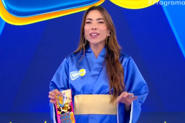 A apresentadora Patricia Abravanel está no palco do programa Silvio Santos. Ela veste um quimono azul com um cinto dourado e segura um bloco da Tele Sena de Primavera. Patrícia sorri e olha para a câmera.
