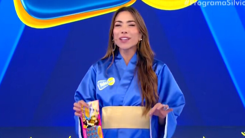A apresentadora Patricia Abravanel está no palco do programa Silvio Santos. Ela veste um quimono azul com um cinto dourado e segura um bloco da Tele Sena de Primavera. Patrícia sorri e olha para a câmera.