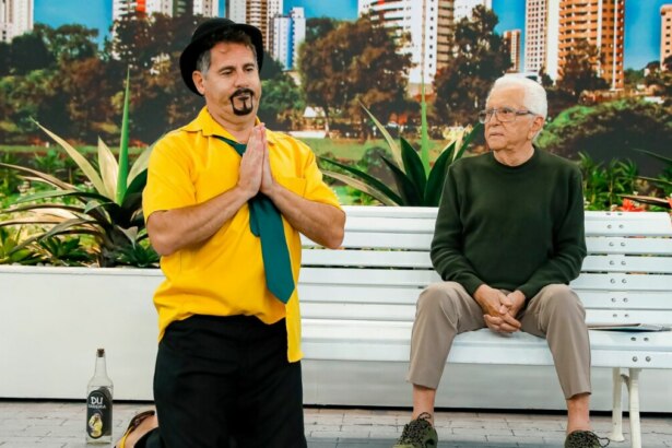 O comediante Saideira, ajoelhado, com as mãos unidas e uma garrafa de cachaça ao lado, e Carlos Alberto de Nóbrega sentados em um banco, em trecho de A Praça é Nossa
