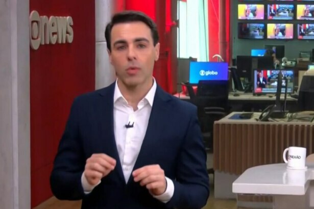 Rafael Colombo, com um terno azul-marinho e camisa branca, está em pé no cenário do estúdio da GloboNews, olhando para a câmera com as mãos juntas.