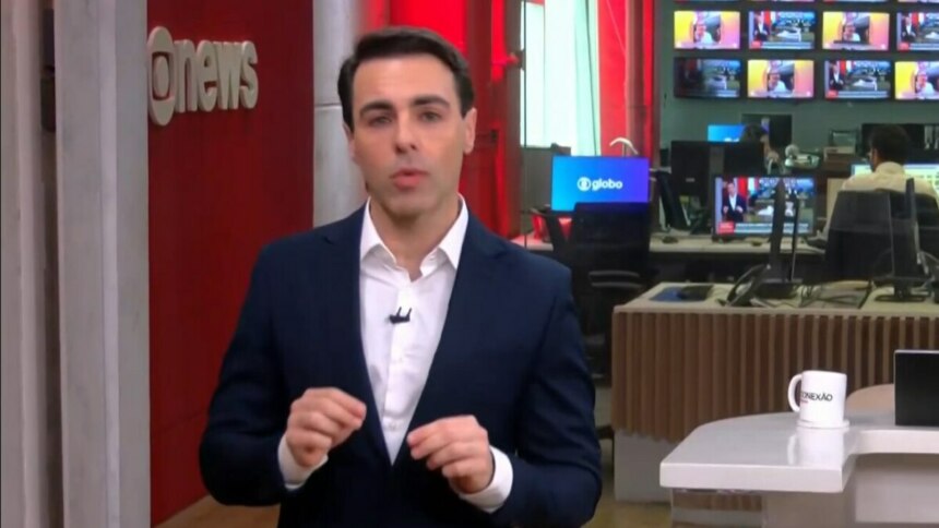 Rafael Colombo, com um terno azul-marinho e camisa branca, está em pé no cenário do estúdio da GloboNews, olhando para a câmera com as mãos juntas.