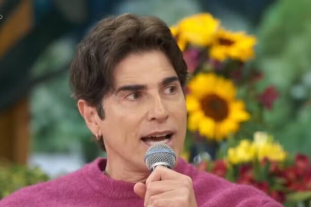 O ator Reynaldo Gianecchini, de suéter rosa, está com um microfone na mão e falando em uma participação no programa Encontro, com um fundo de arranjos de flores.