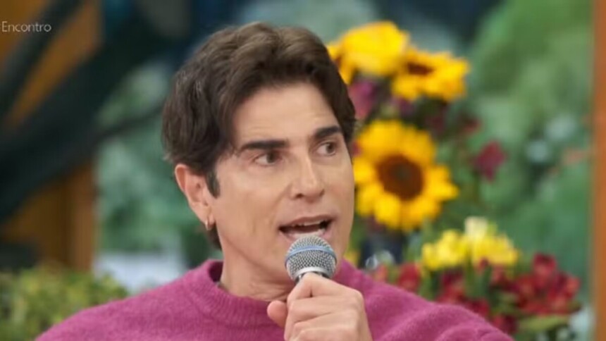 O ator Reynaldo Gianecchini, de suéter rosa, está com um microfone na mão e falando em uma participação no programa Encontro, com um fundo de arranjos de flores.