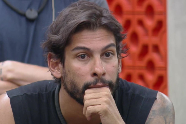 O participante Shia de blusa preta, com barba e cabelos longos, olha para o lado com uma expressão séria, em uma cena do reality show A Fazenda.
