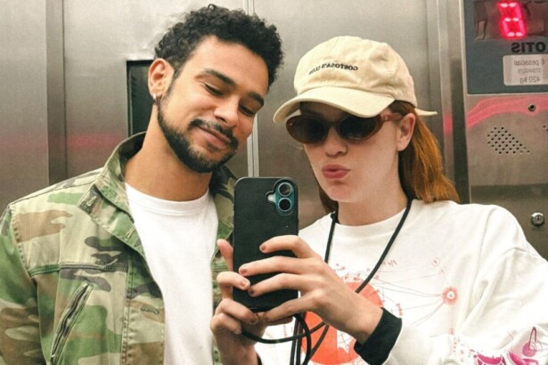 O ator Sérgio Malheiros, de jaqueta camuflada, e a atriz Sophia Abrahão, de boné bege e óculos escuros, fazem uma selfie juntos dentro de um elevador. Ele está com os olhos fechados e a abraça, enquanto ela segura o celular e faz bico.