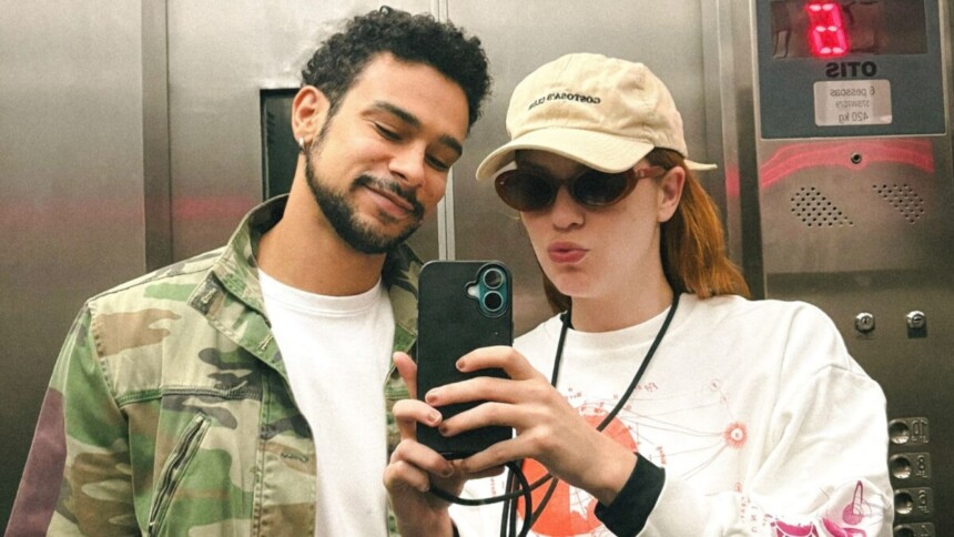 O ator Sérgio Malheiros, de jaqueta camuflada, e a atriz Sophia Abrahão, de boné bege e óculos escuros, fazem uma selfie juntos dentro de um elevador. Ele está com os olhos fechados e a abraça, enquanto ela segura o celular e faz bico.