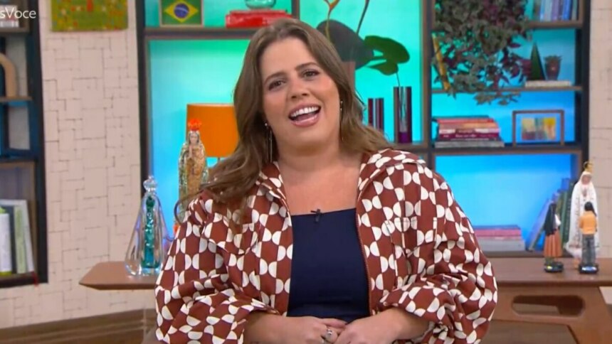 Tati Machado sorrindo para a câmera, usando uma jaqueta estampada e uma blusa azul-marinho, sentada no cenário colorido e aconchegante do programa Mais Você.