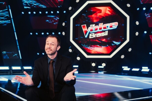 O apresentador Tiago Leifert, de terno e gravata, está em um palco escuro com a logomarca do The Voice Brasil em neon ao fundo. Ele está agachado com as mãos para o alto, sorrindo e olhando para a câmera.