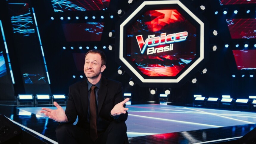 O apresentador Tiago Leifert, de terno e gravata, está em um palco escuro com a logomarca do The Voice Brasil em neon ao fundo. Ele está agachado com as mãos para o alto, sorrindo e olhando para a câmera.