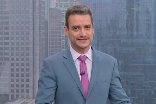 O jornalista Tiago Scheuer está em um estúdio de TV no Bom Dia SP, vestido com um terno azul claro, camisa branca e gravata rosa. O fundo é um cenário de cidade.