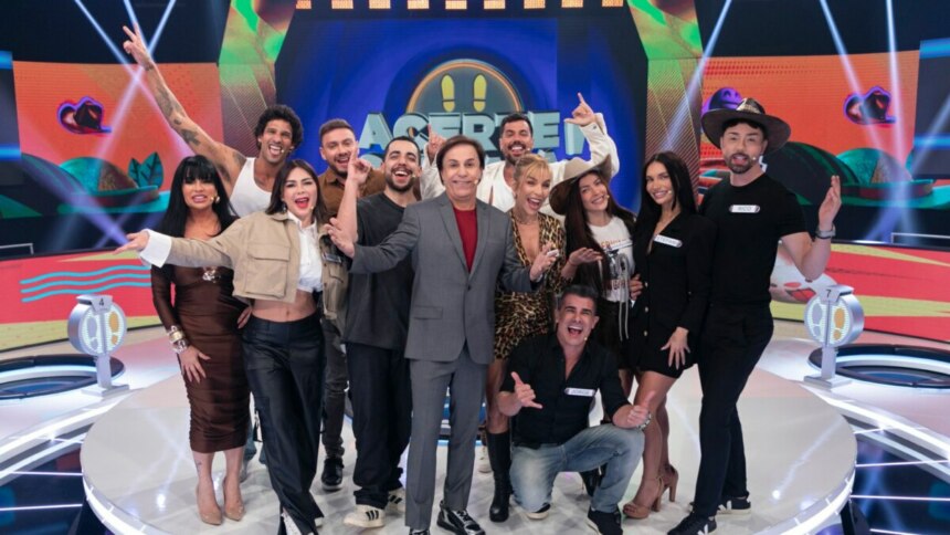 Tom Cavalcante com um terno cinza posa com os 12 participantes do reality show "Acerte ou Caia!" no palco, todos sorrindo e em clima de festa.