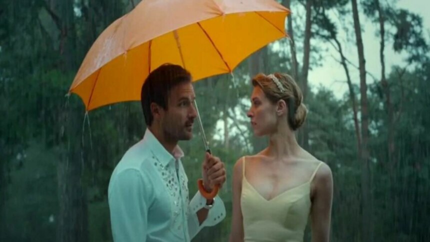 Cena do filme Uma Noiva em Apuros. Um casal está na chuva, sob um guarda-chuva laranja. Eles se olham intensamente em um momento romântico.