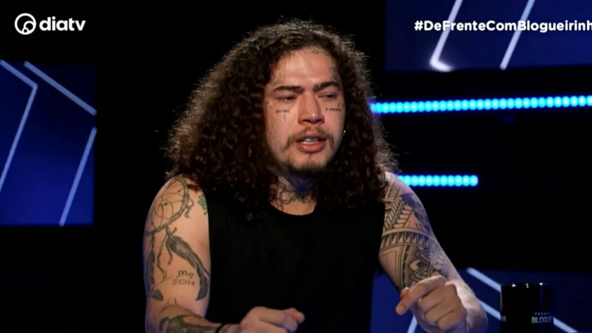 Whindersson Nunes, de cabelos cacheados e longos, com tatuagens no rosto e nos braços, olha para baixo e tem expressão emocionada no programa De Frente com Blogueirinha.