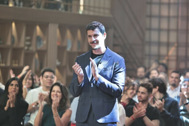 Felipe B. batendo palmas no centro do cenário do MasterChef Brasil 2025. Atrás dele um aplateia também bate palmas