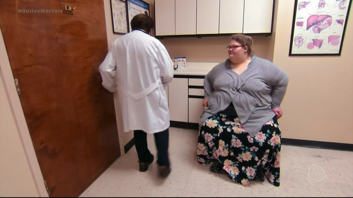 Seana Collins em consulta médica, sentada em sala de exame com médico ao fundo.
