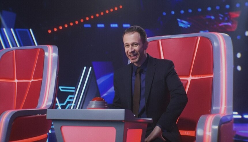 Tiago Leifert sentado em uma cadeira do The Voice Brasil 2025 e sorridente