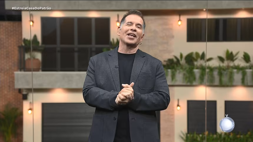 Leandro Hassum com as mãos unidas e sorridente no centro do cenário estúdio glass. Ao fundo a Casa do Patrão