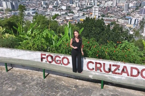 Camila Busnello em cima de um morro, com uma frase digitalizada escrito Fogo Cruzado no muro em chamada do Domingo Espetacular